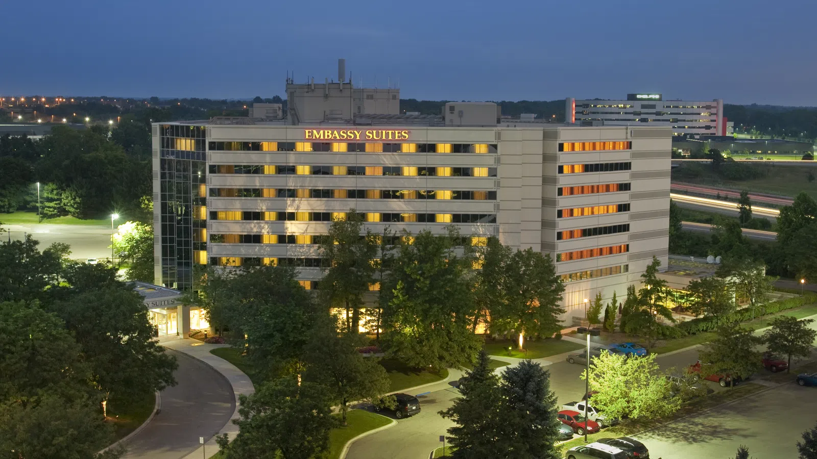Night view of Embassy Suites hotel building with illuminated windows and surrounding trees and parking lot.