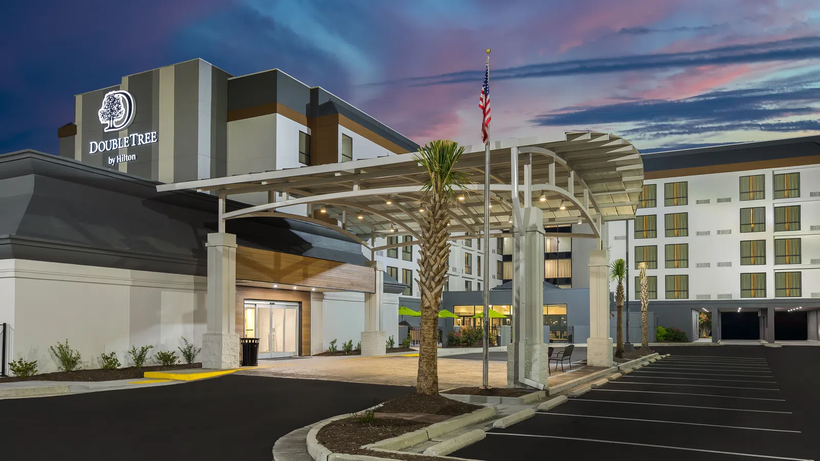 DoubleTree by Hilton hotel exterior at dusk with lit entrance, palm trees, and American flag in clear sky.