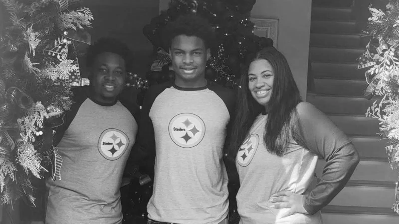 Three people wearing matching Steelers shirts and plaid pajama pants standing by Christmas decorations indoors