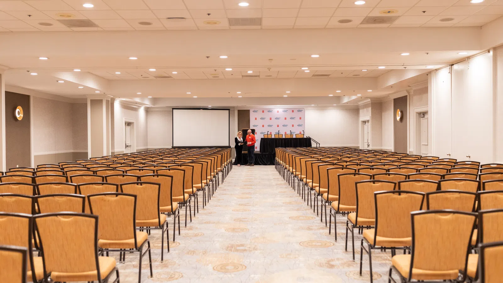 Empty conference room with rows of chairs facing a stage and projector screen under ceiling lights.