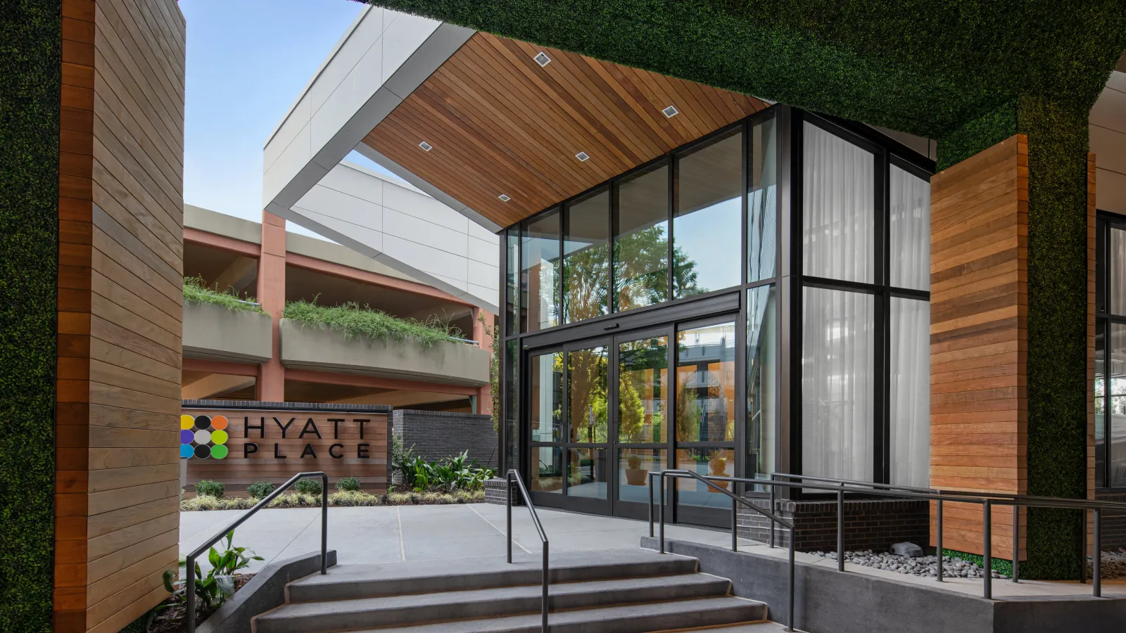Entrance of Hyatt Place hotel with glass doors, wooden accents, greenery, and modern architectural design.