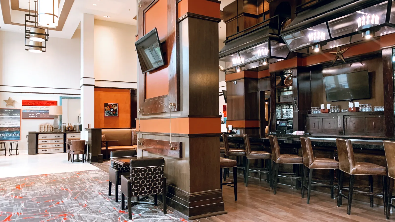 Spacious hotel lounge with wooden bar, high stools, patterned carpet, and modern pendant lighting.