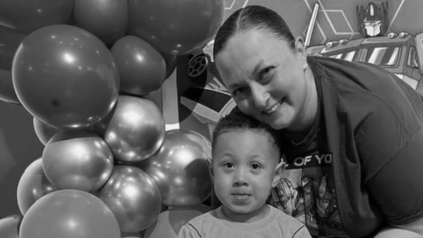 Child and adult smiling in front of birthday balloons and happy birthday banner