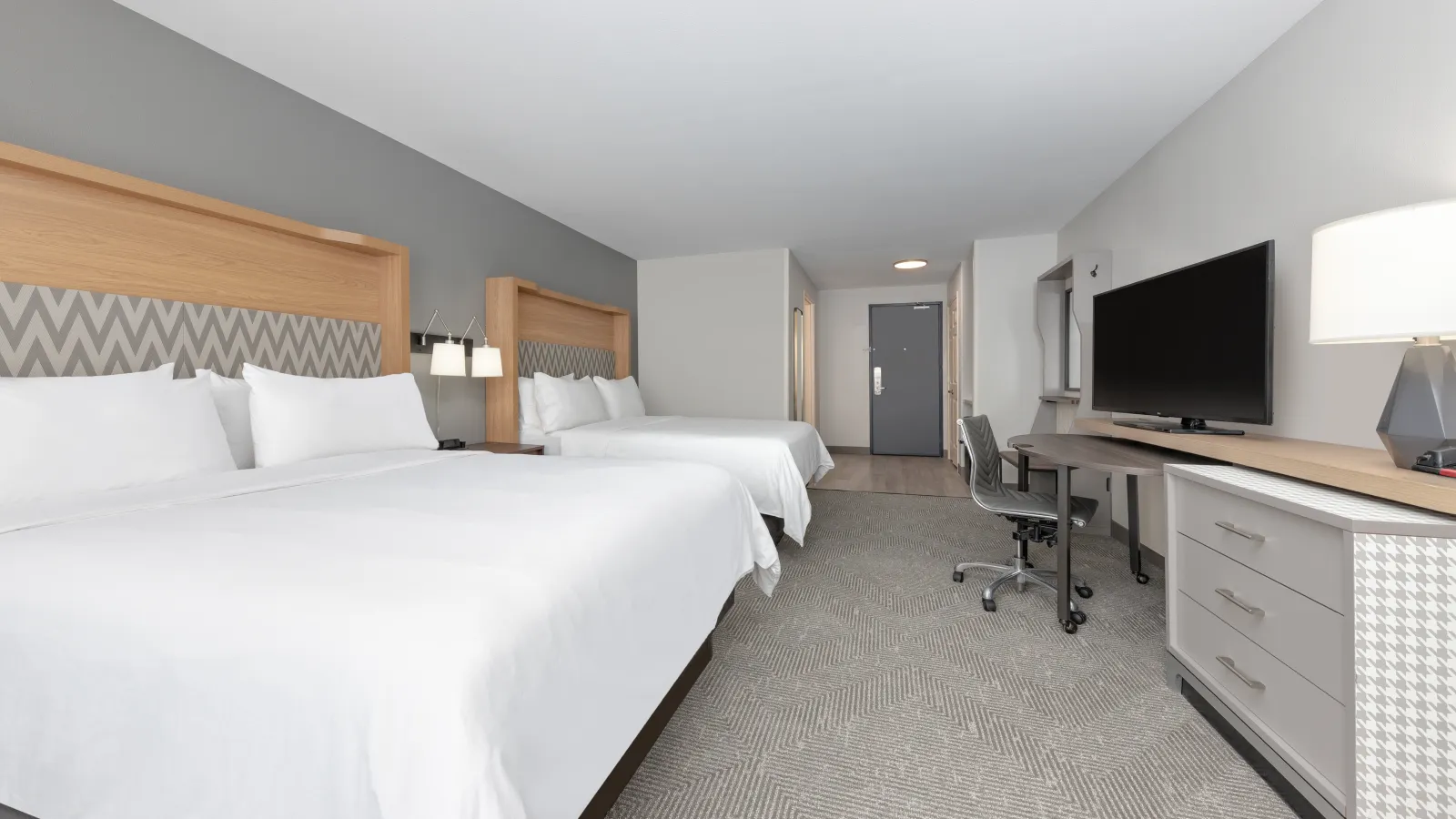 Modern hotel room with two queen beds, gray decor, flat-screen TV, desk, and cozy lighting