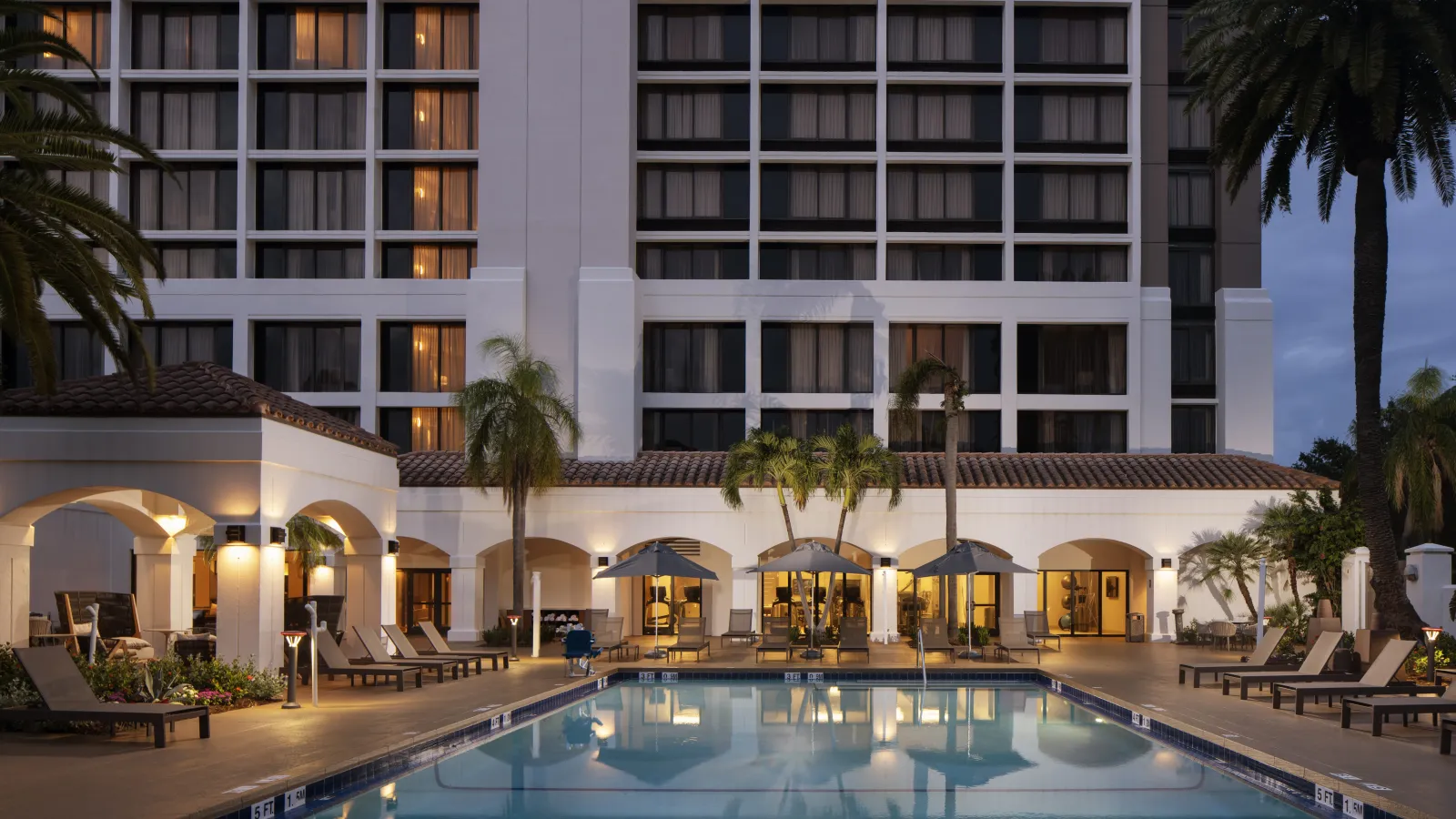 Evening view of a hotel pool area with lounge chairs, umbrellas, palm trees, and illuminated building windows.