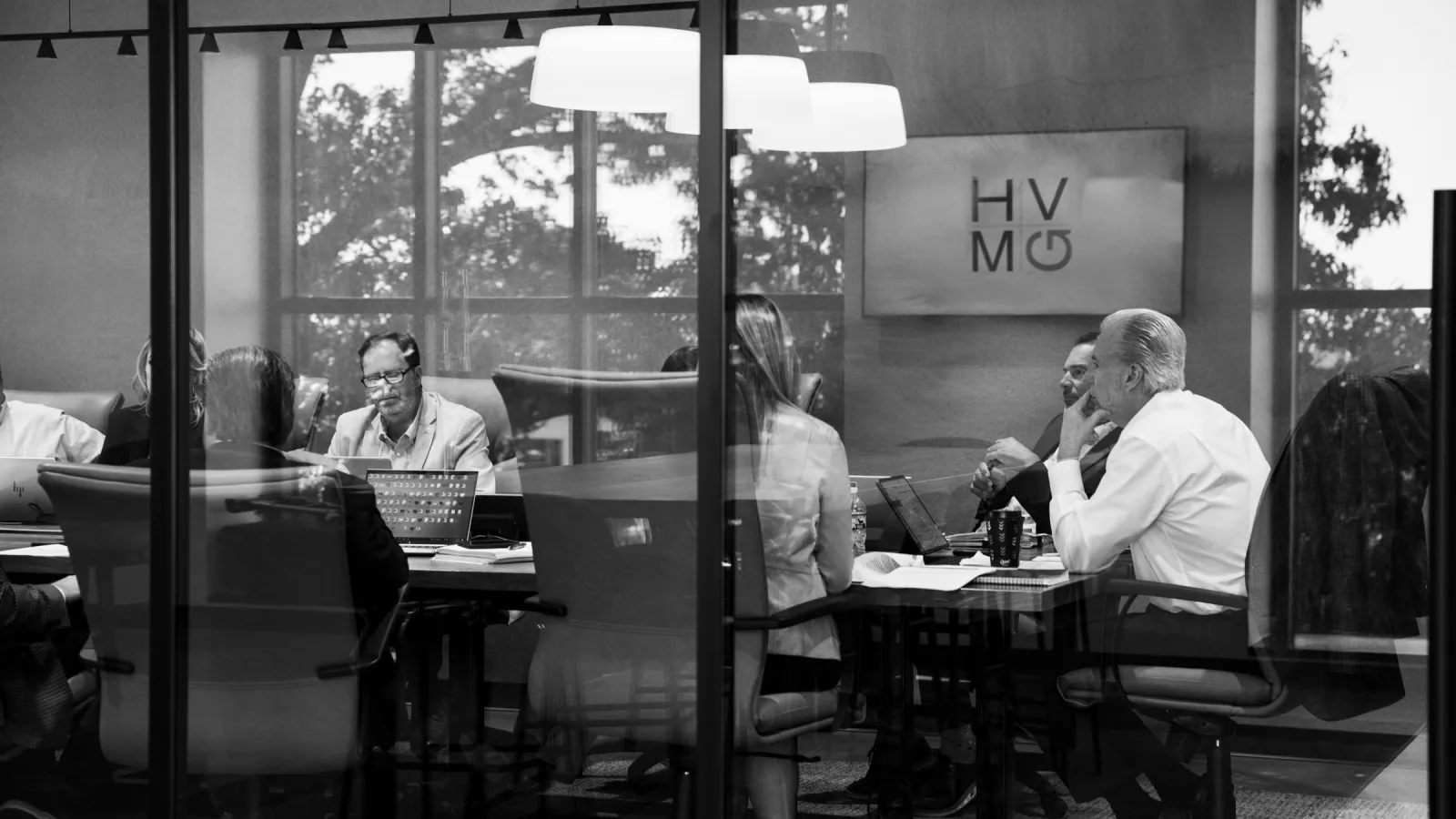 Black and white photo of business professionals engaged in a meeting inside a modern glass-walled conference room.