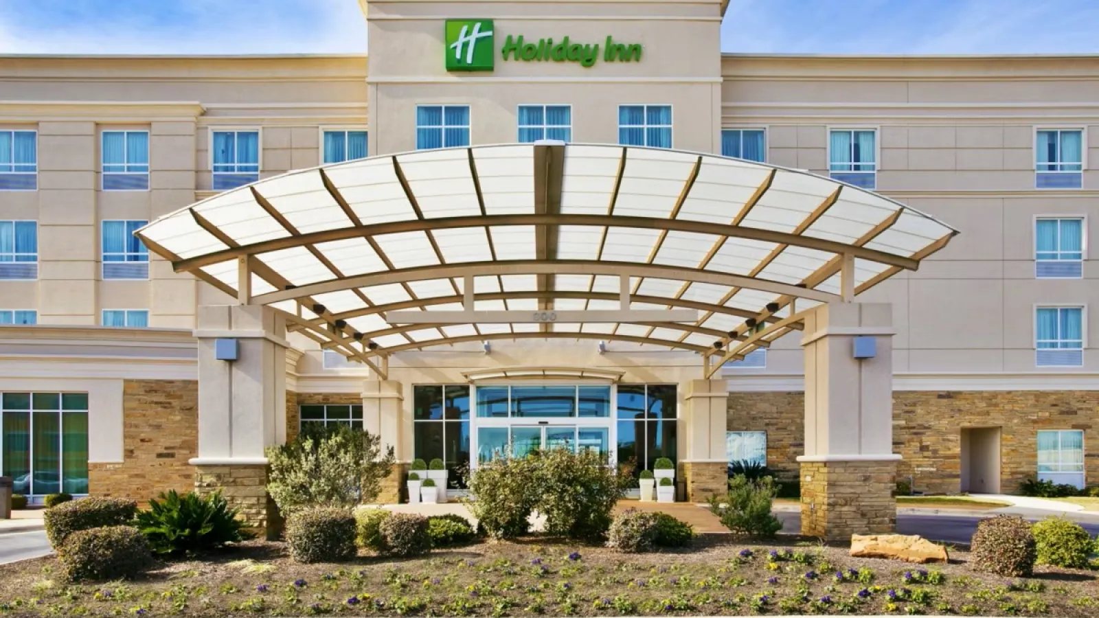 Front entrance of a Holiday Inn hotel with glass canopy, landscaping, and clear blue sky background.