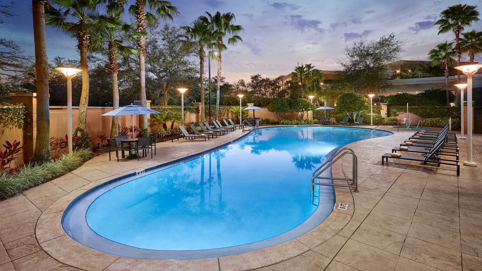Outdoor hotel pool at dusk surrounded by palm trees, lounge chairs, and lit street lamps under a purple sky