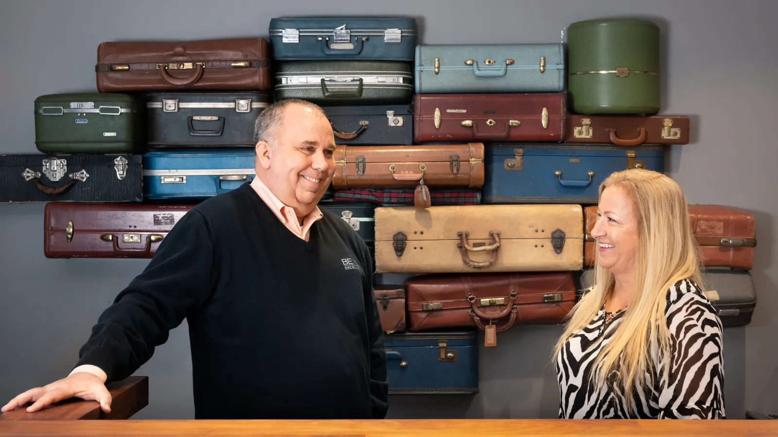 a man and a woman sitting at a desk with a large stack of suitcases