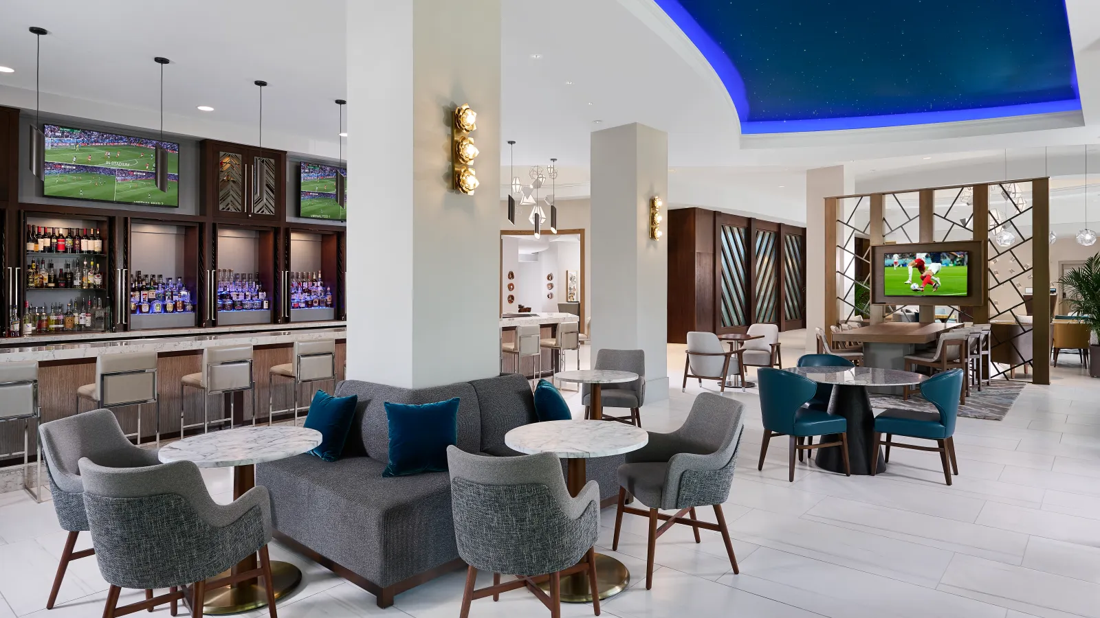 Modern lounge area with cozy seating, marble tables, bar counter, and multiple TVs displaying sports events.