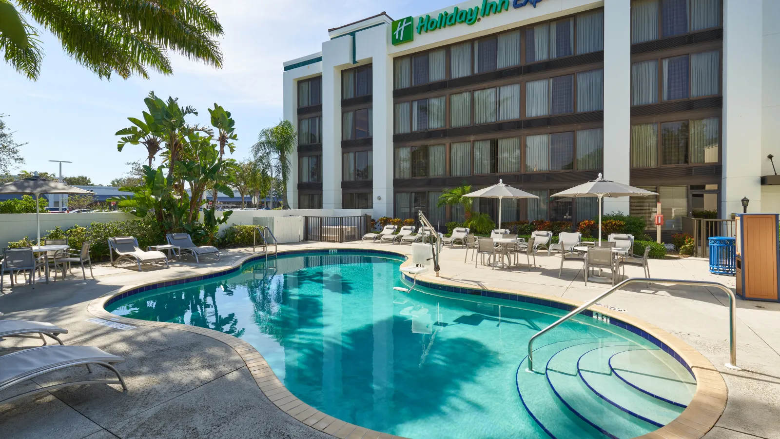 Outdoor swimming pool at Holiday Inn Express with lounge chairs, umbrellas, and palm trees on a sunny day.