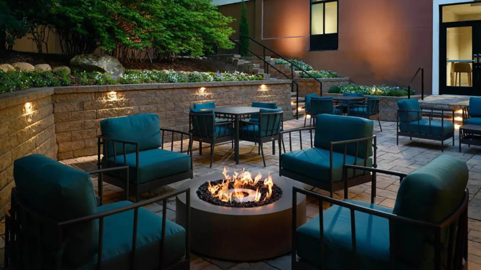 Cozy outdoor patio with teal cushioned chairs around a lit fire pit and tables in the background at dusk.