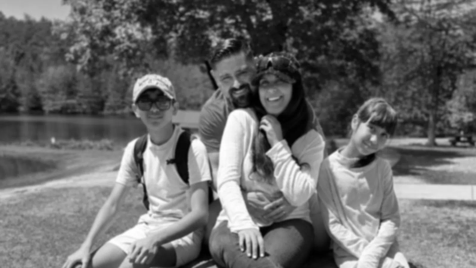 Family of four sitting on a large rock in a park by a lake on a sunny day.