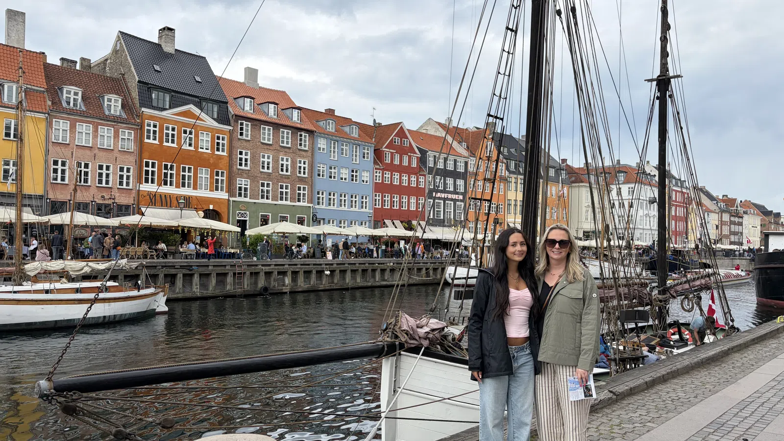 Two women standing by a canal with colorful historic buildings and boats in the background under a cloudy sky.