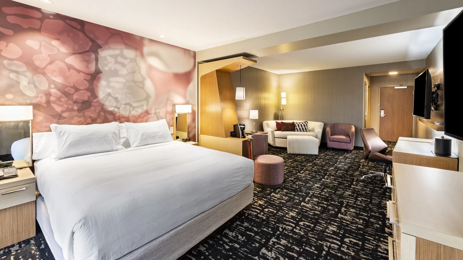 Modern hotel room with king-size bed, abstract pink wall art, seating area, workspace, and neutral decor.