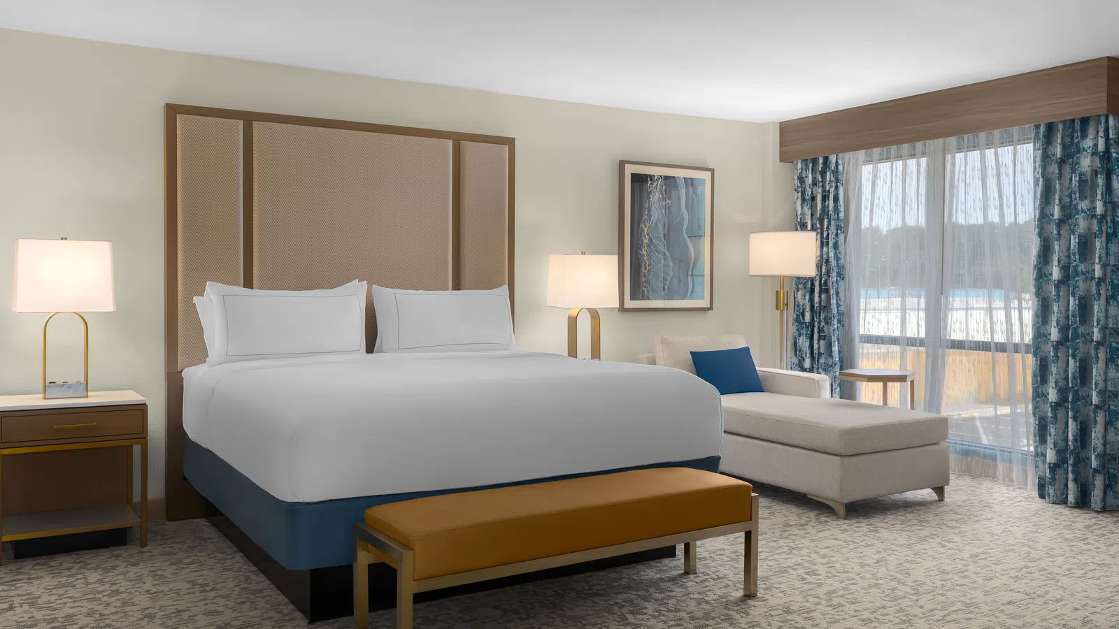 Modern hotel room with king bed, beige chaise lounge, wooden bench, lamps, and balcony with patterned curtains.