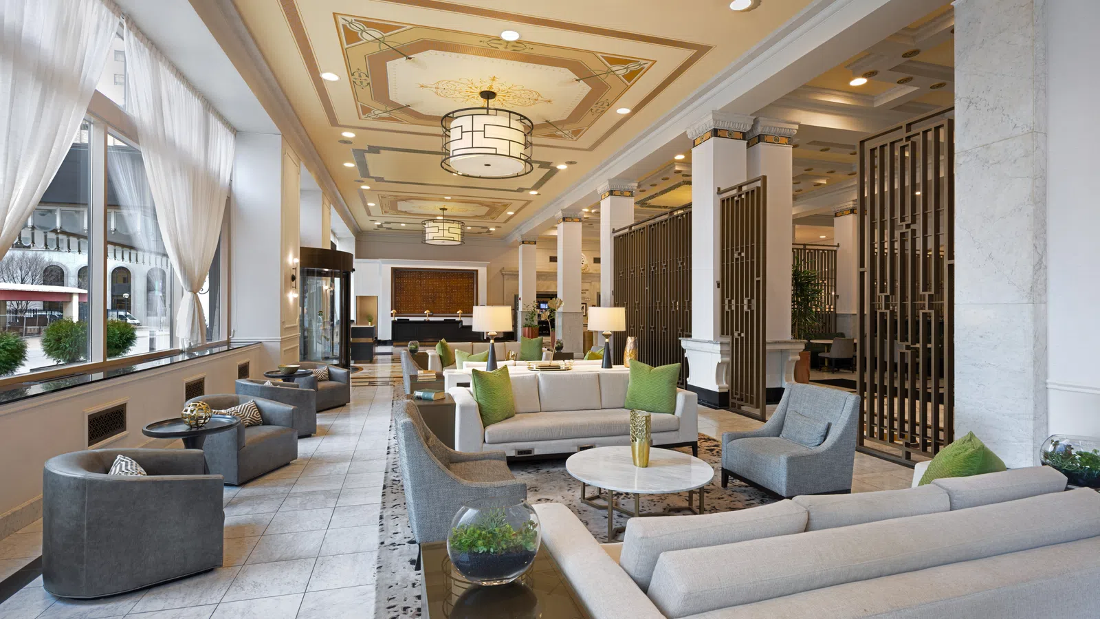 Bright modern hotel lobby with grey and cream sofas, green cushions, large windows, and decorative ceiling lights.