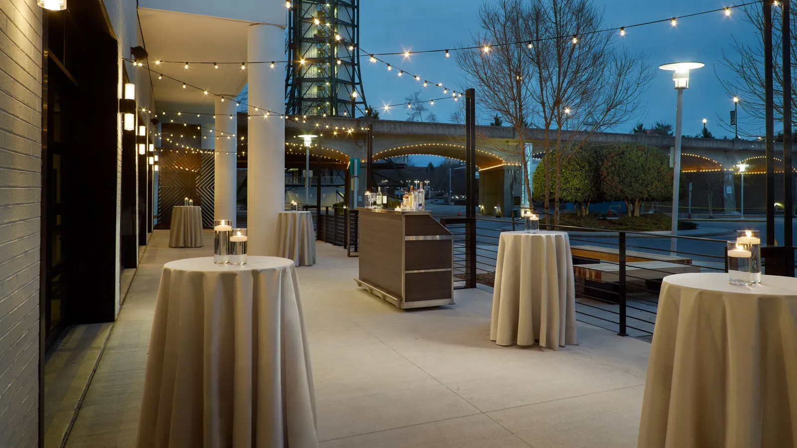 Outdoor evening event space with round cocktail tables, string lights, and a waterfront view under a bridge.