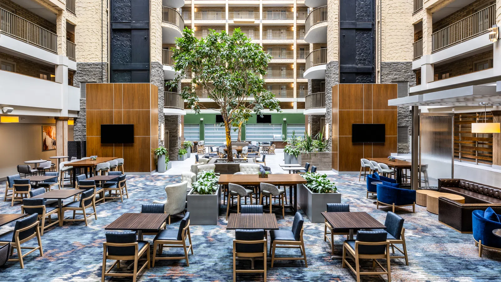 Modern hotel lobby with seating areas, large central tree, wood accents, and multi-story balcony views