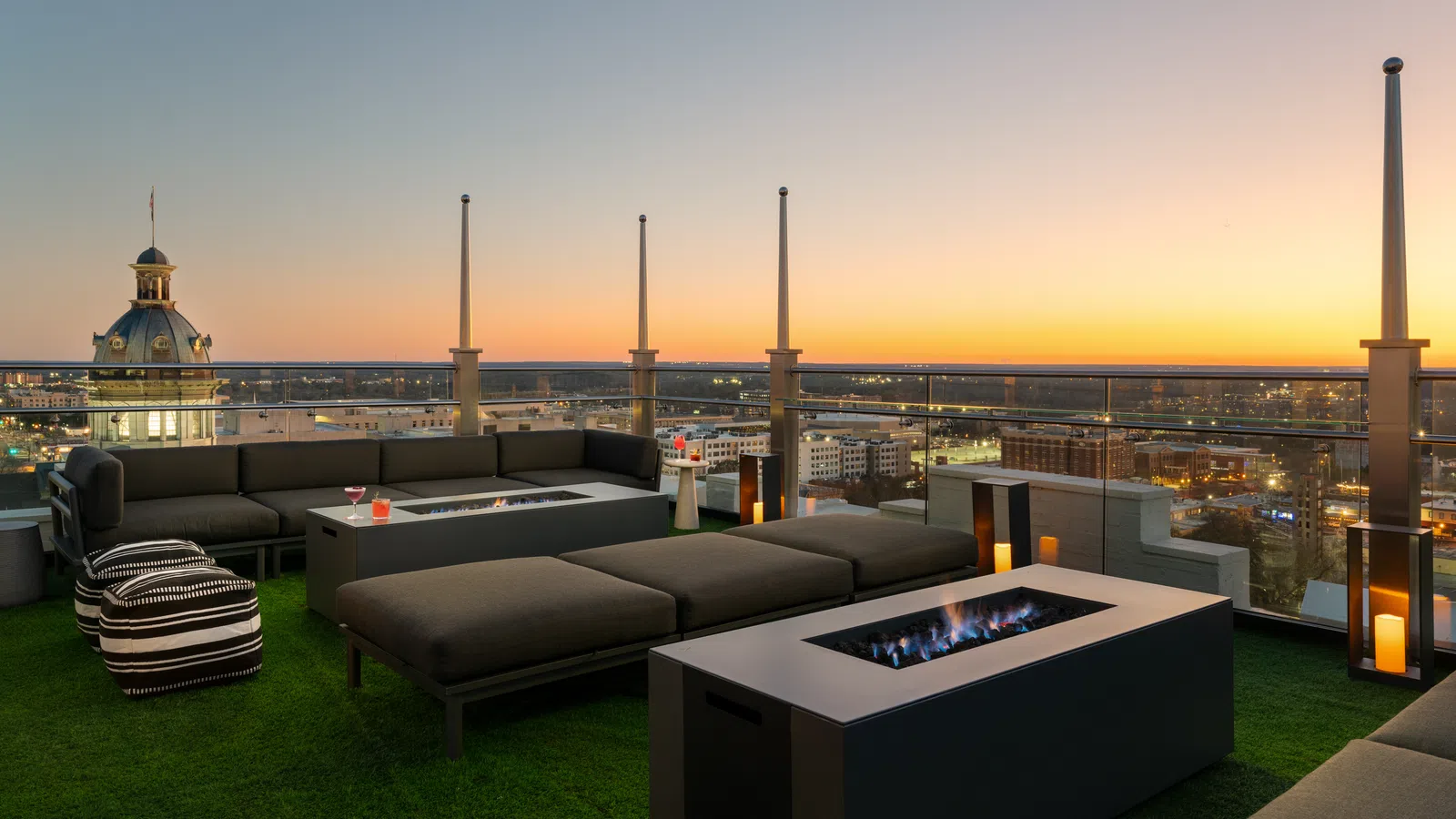 Modern rooftop lounge with fire pits, comfortable sofas, and city skyline at sunset with clear sky.