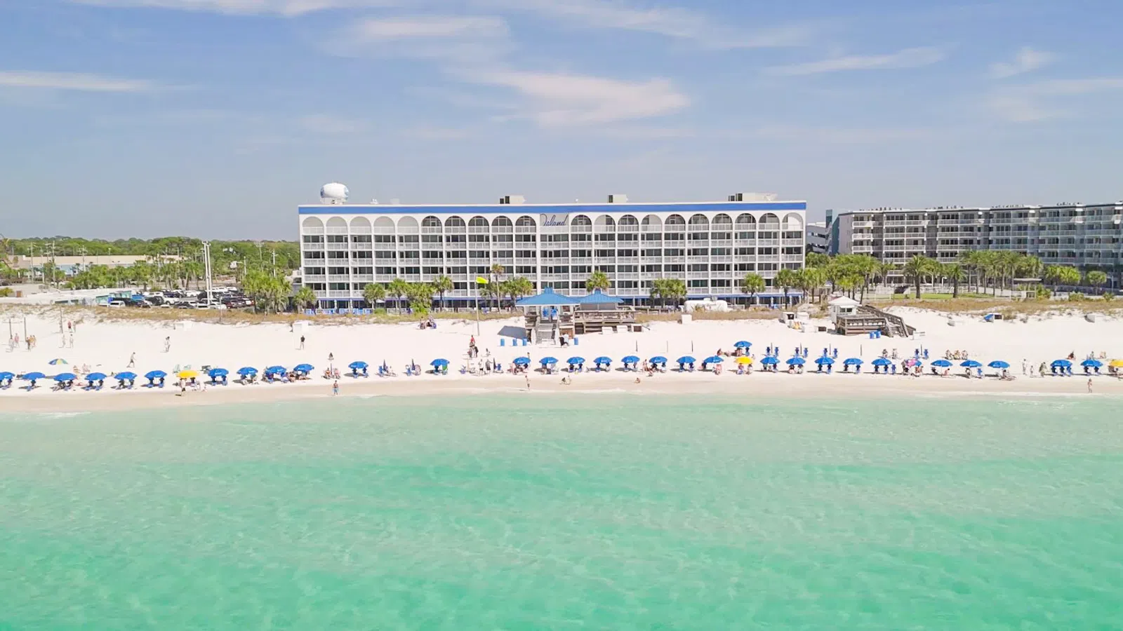A beachfront hotel with rows of blue umbrellas on white sand and turquoise ocean water under a clear sky.