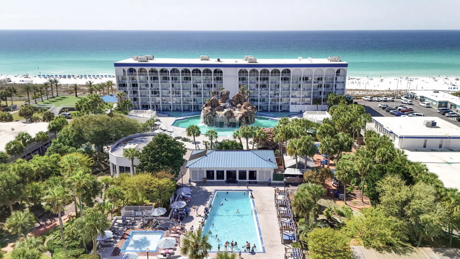 A beachfront hotel with a large pool, rock waterfall, palm trees, and turquoise ocean in the background under a sunny sky