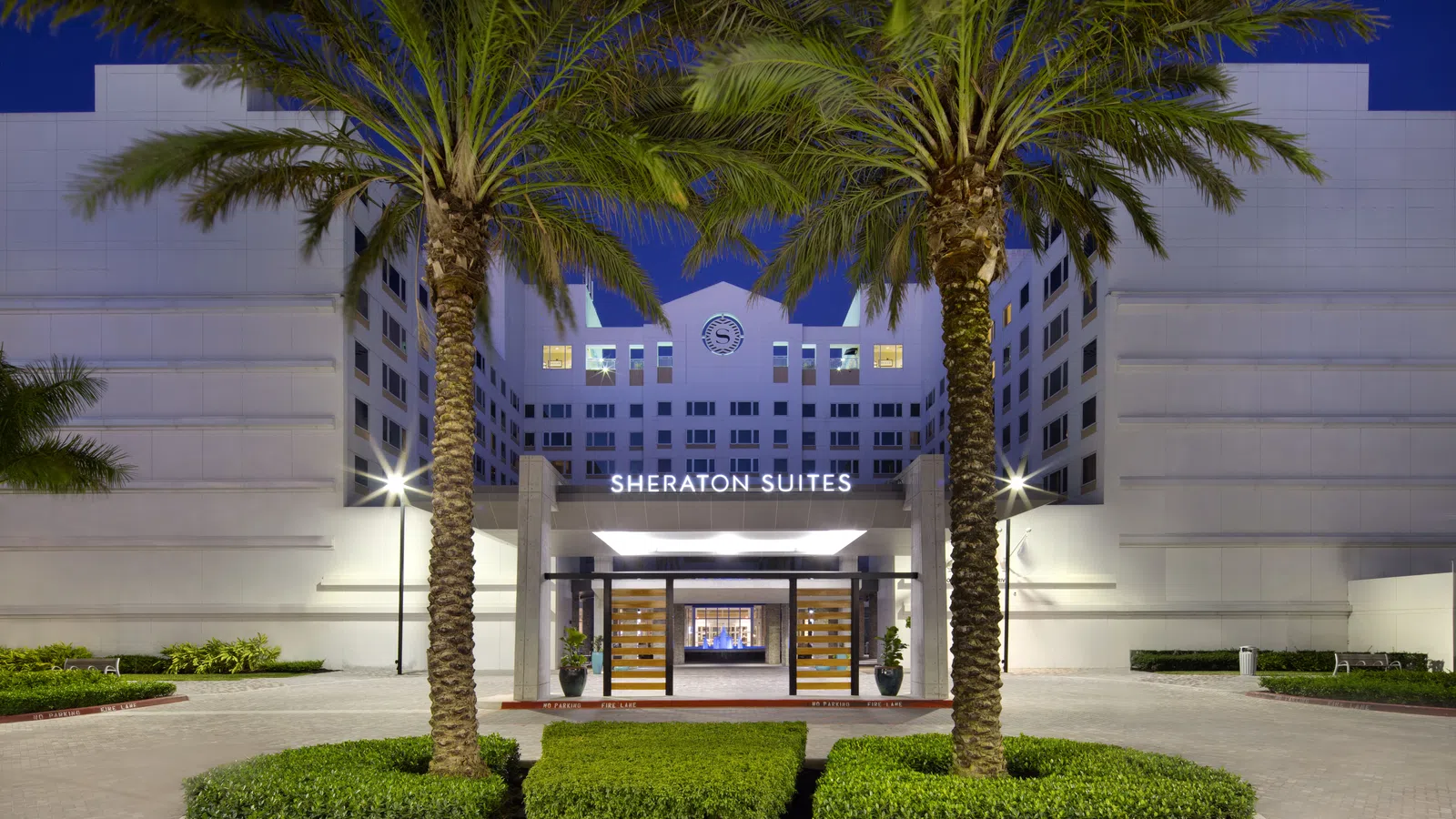 Exterior view of Sheraton Suites hotel entrance framed by palm trees and illuminated at dusk.