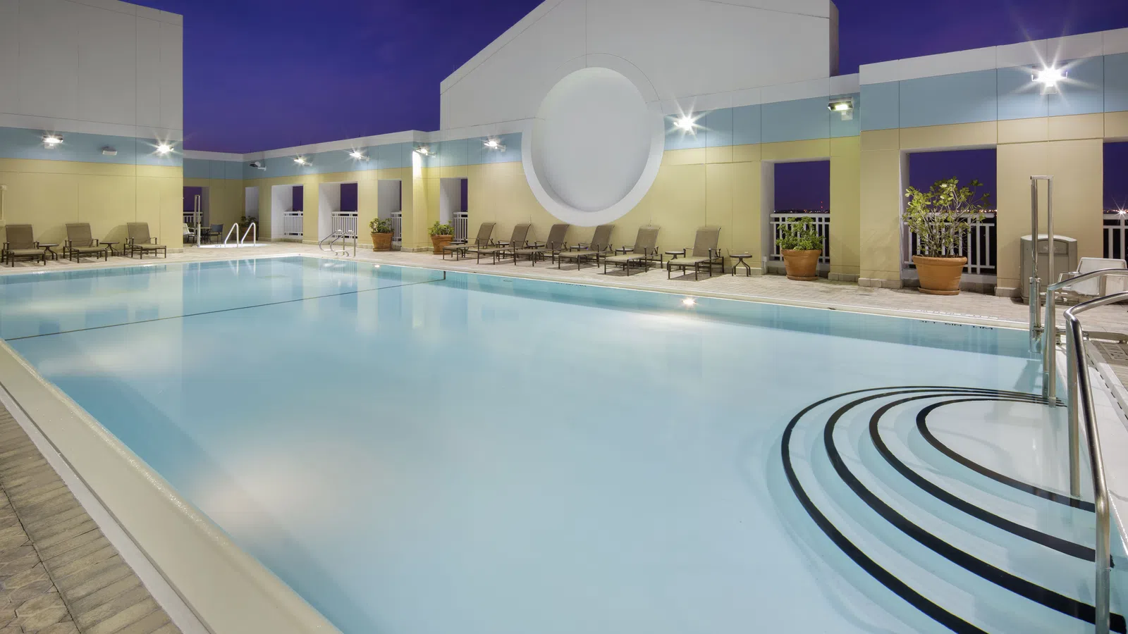 Modern rooftop swimming pool with lounge chairs and potted plants under evening sky at a hotel or apartment.