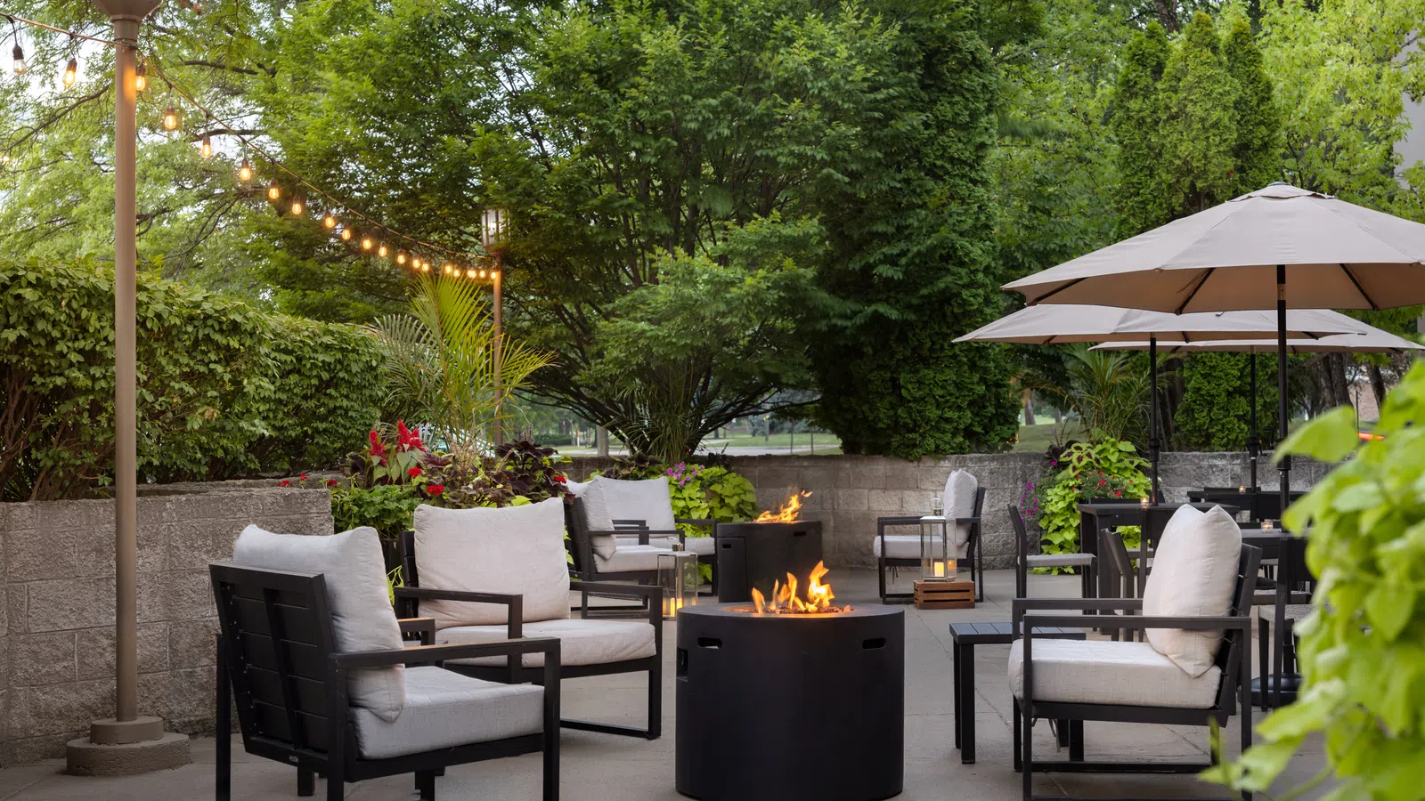 Cozy outdoor patio with cushioned chairs, fire pits, string lights, umbrellas, and lush greenery in the evening.