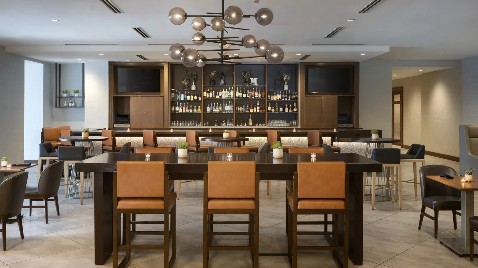 Modern hotel bar with high tables, leather chairs, ambient lighting, and a well-stocked backlit liquor display.