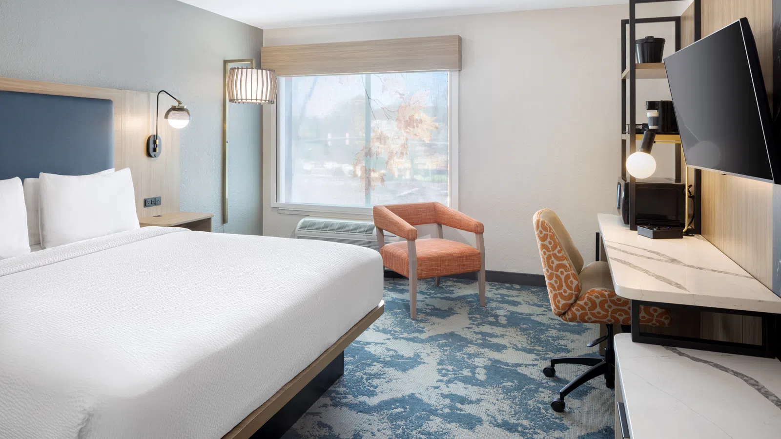 Modern hotel room with king bed, orange chair, patterned desk chair, blue and gray carpet, and large window with shades.