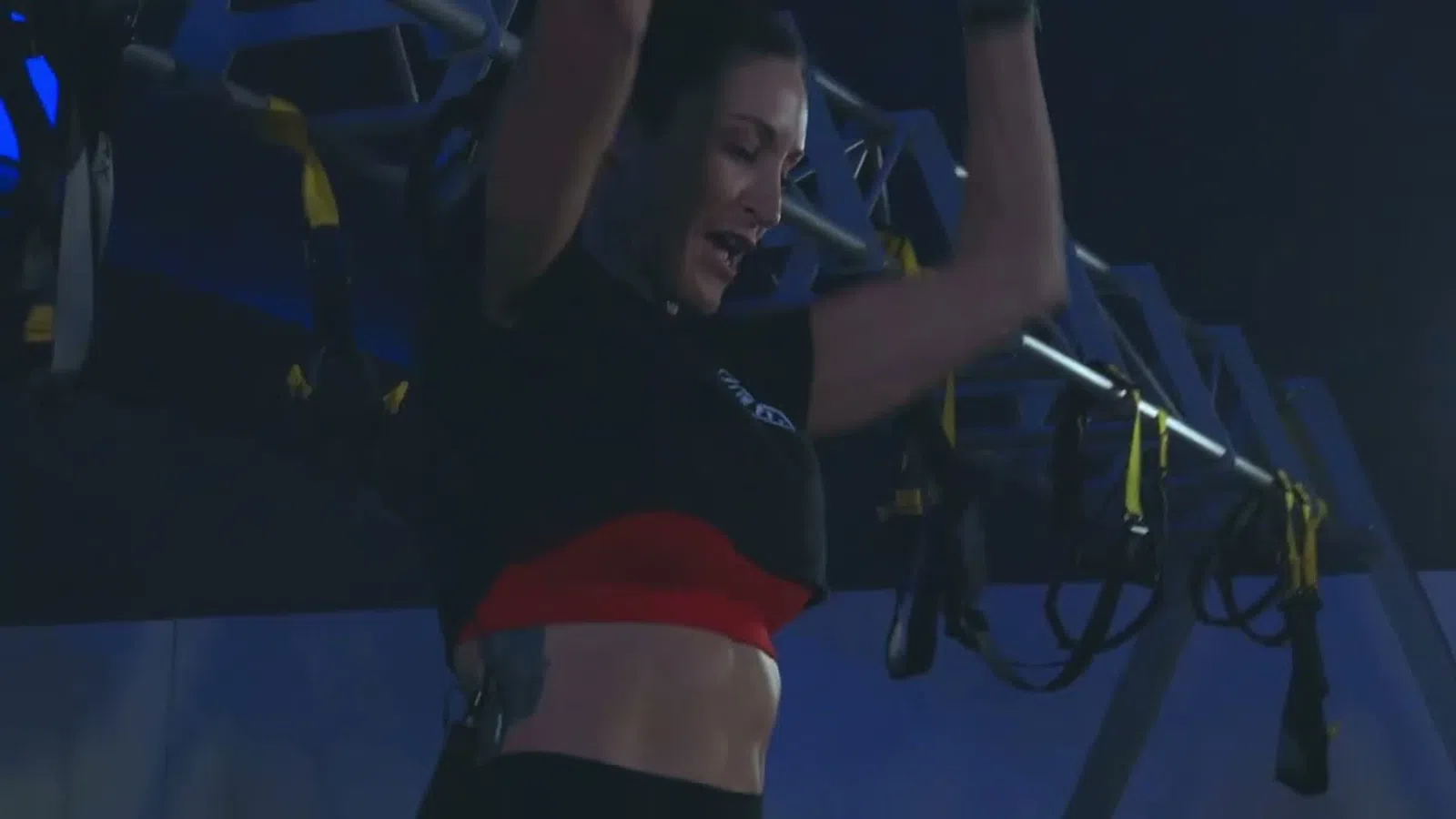 Woman performing pull-ups on gym equipment under dim blue lighting wearing athletic clothing.