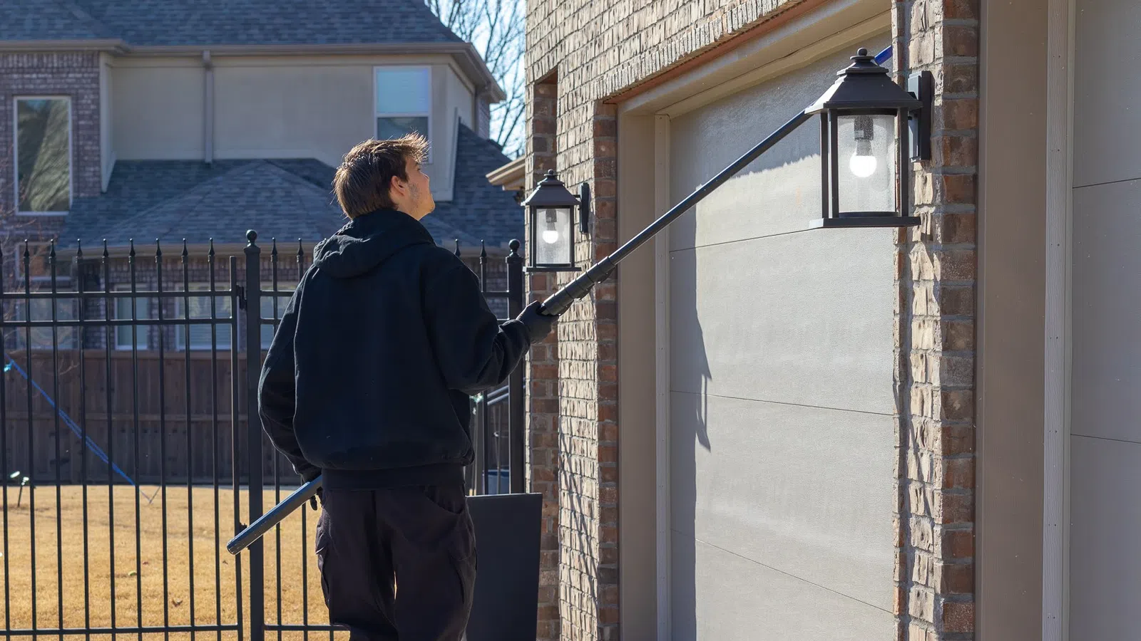 Man in black hoodie using a long pole to change an outdoor light near a garage door in a residential area.