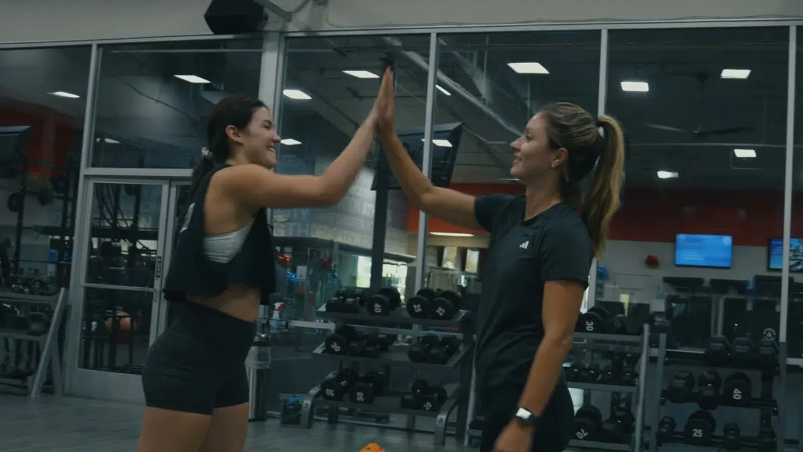 Two women high-fiving in a gym with weights and mirrors in the background during a workout session