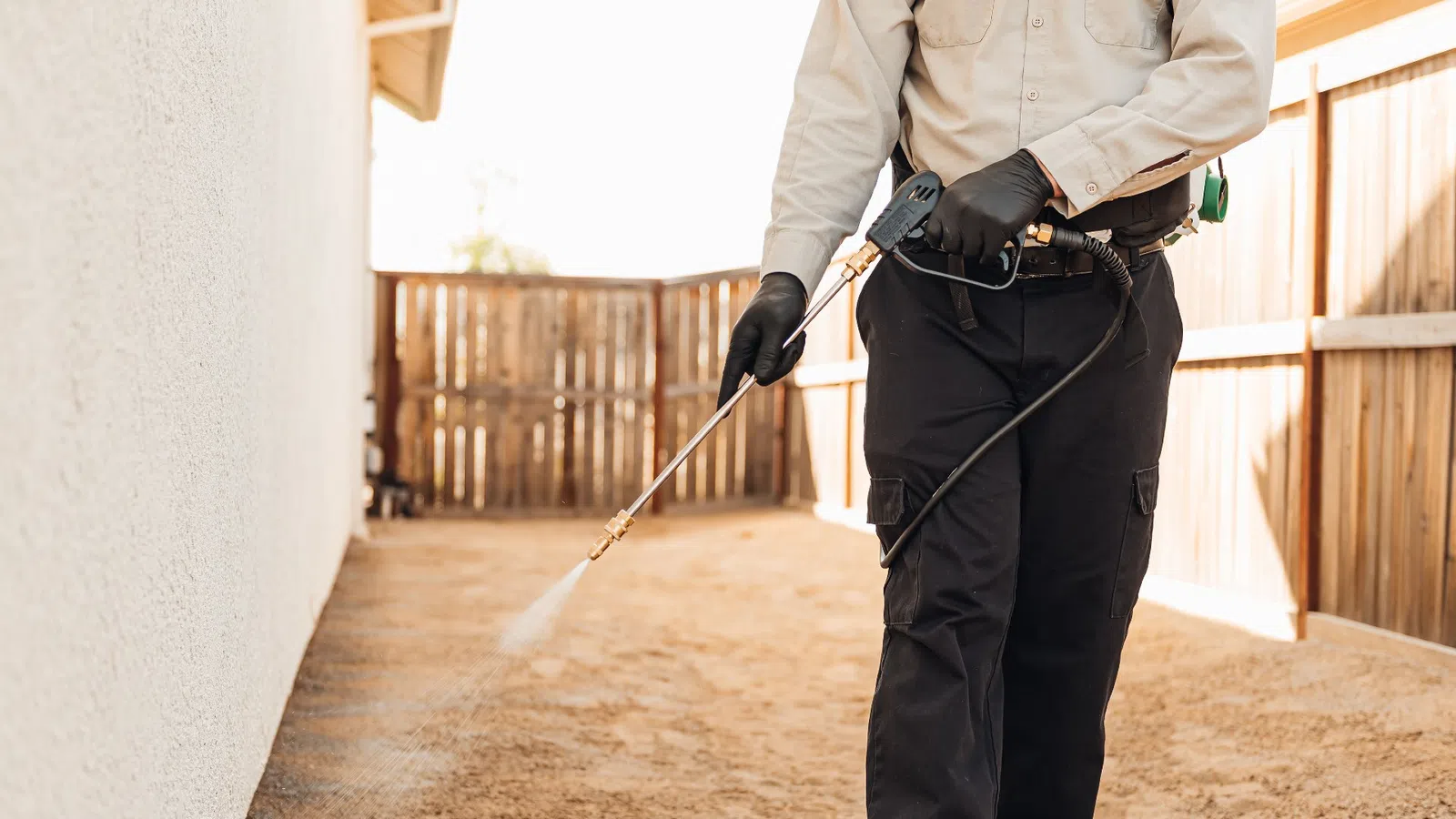 Pest control technician spraying insecticide outside a house near wooden fence on dirt ground.