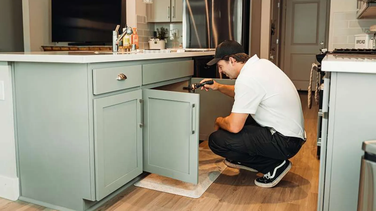 Man inspecting under kitchen sink cabinet with flashlight in modern kitchen with wooden flooring and appliances