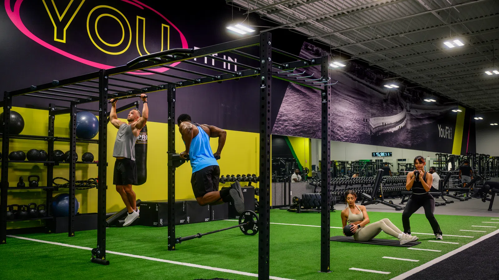 Two men performing pull-ups and ring dips on gym equipment while two women exercise on mat in a modern fitness center.
