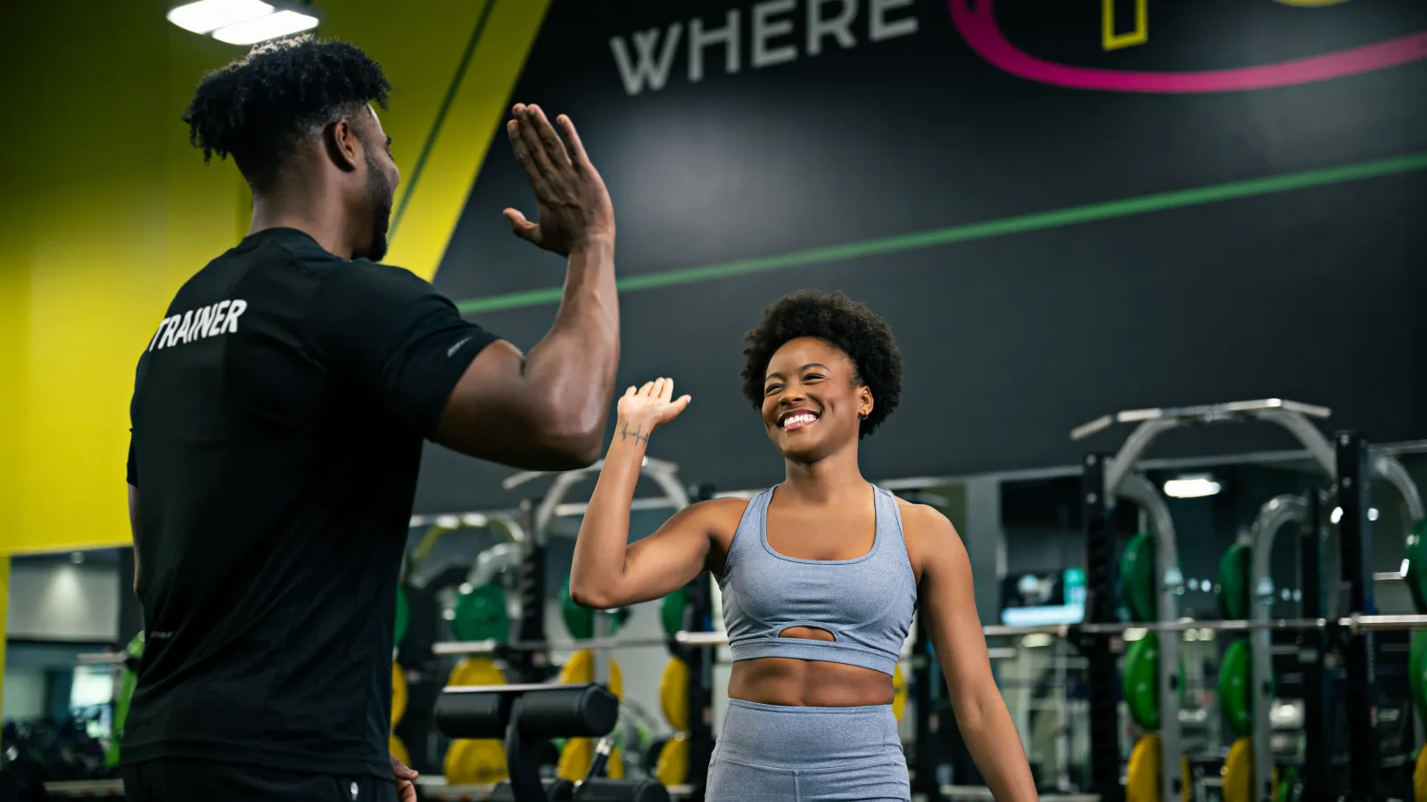 Woman in athletic wear high-fiving her male trainer in a colorful gym setting with weightlifting equipment.