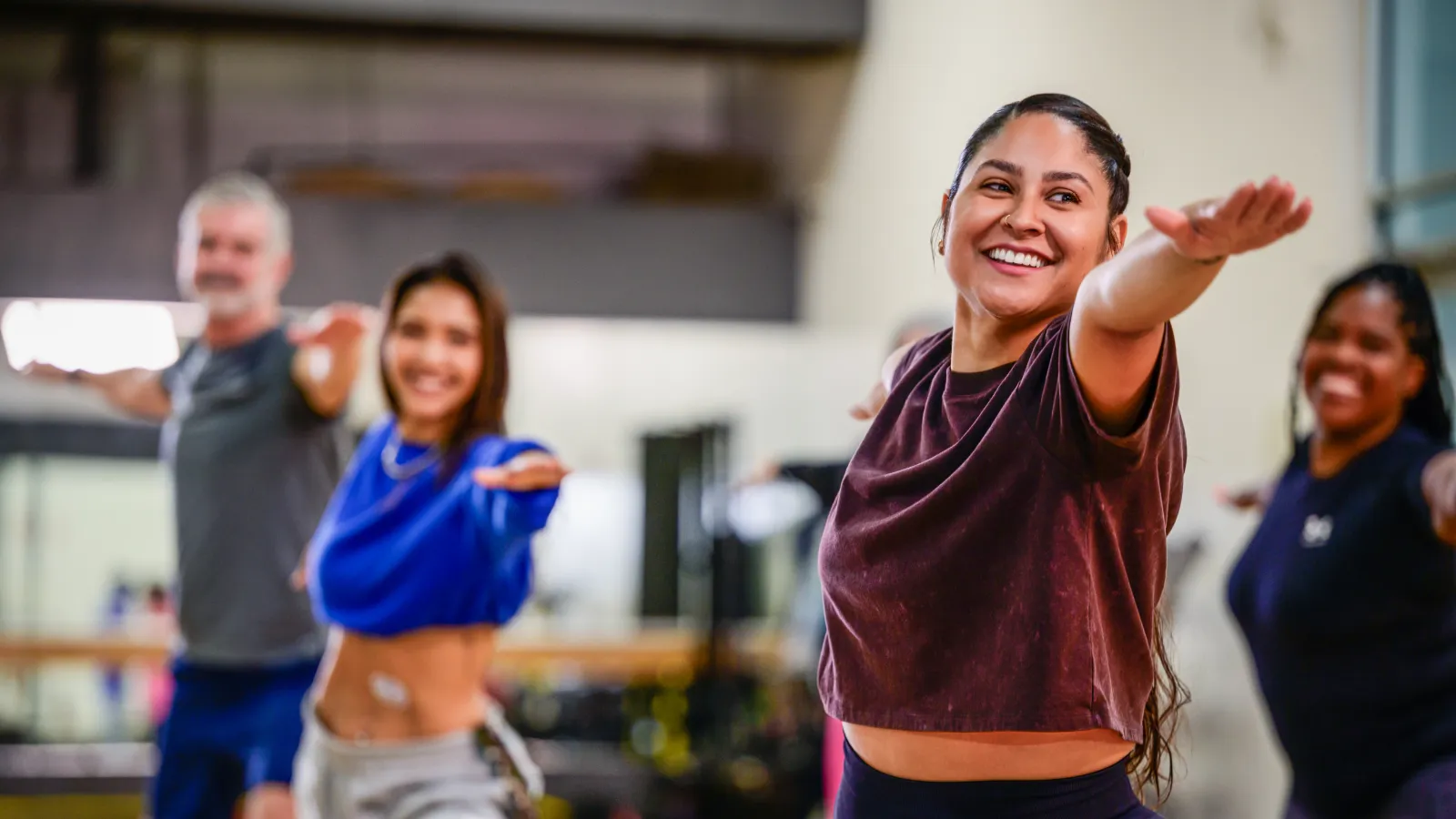 A diverse group of people smiling and stretching their arms forward during a fitness or yoga class indoors.