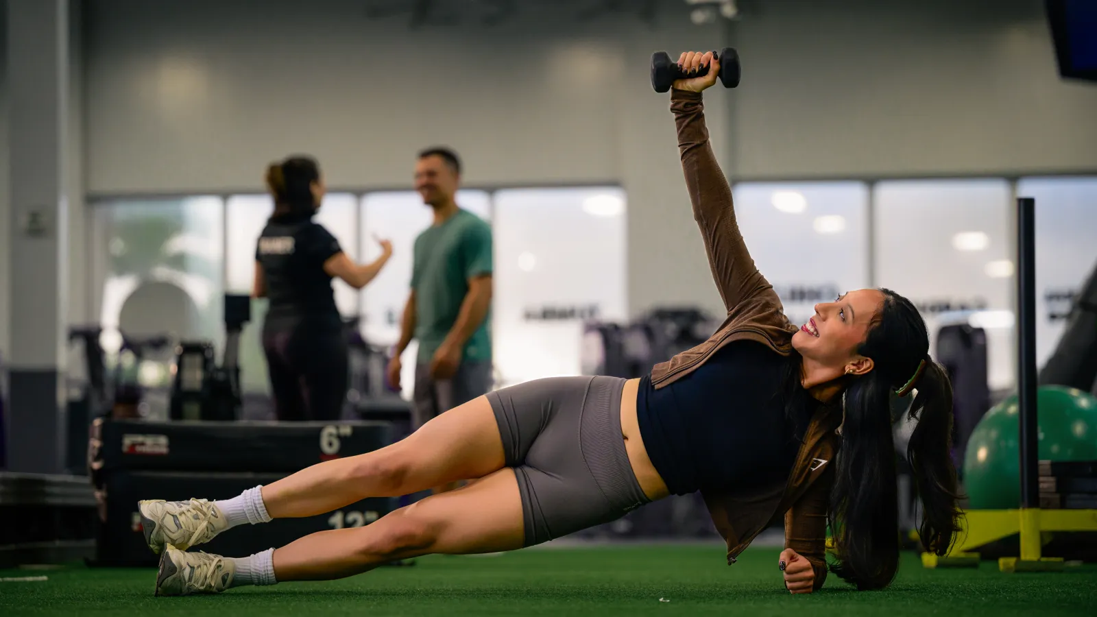 Young woman performs side plank with dumbbell in gym, two people chat in background near workout equipment.