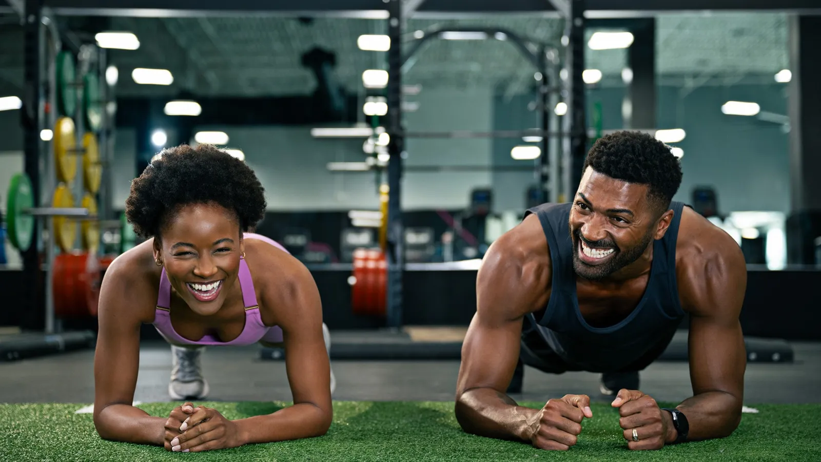 Happy man and woman doing plank exercise together on gym turf with workout equipment in background