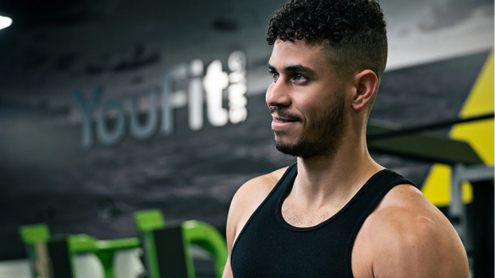 Muscular man in black tank top lifting dumbbell at YouFit gym with green workout equipment in the background