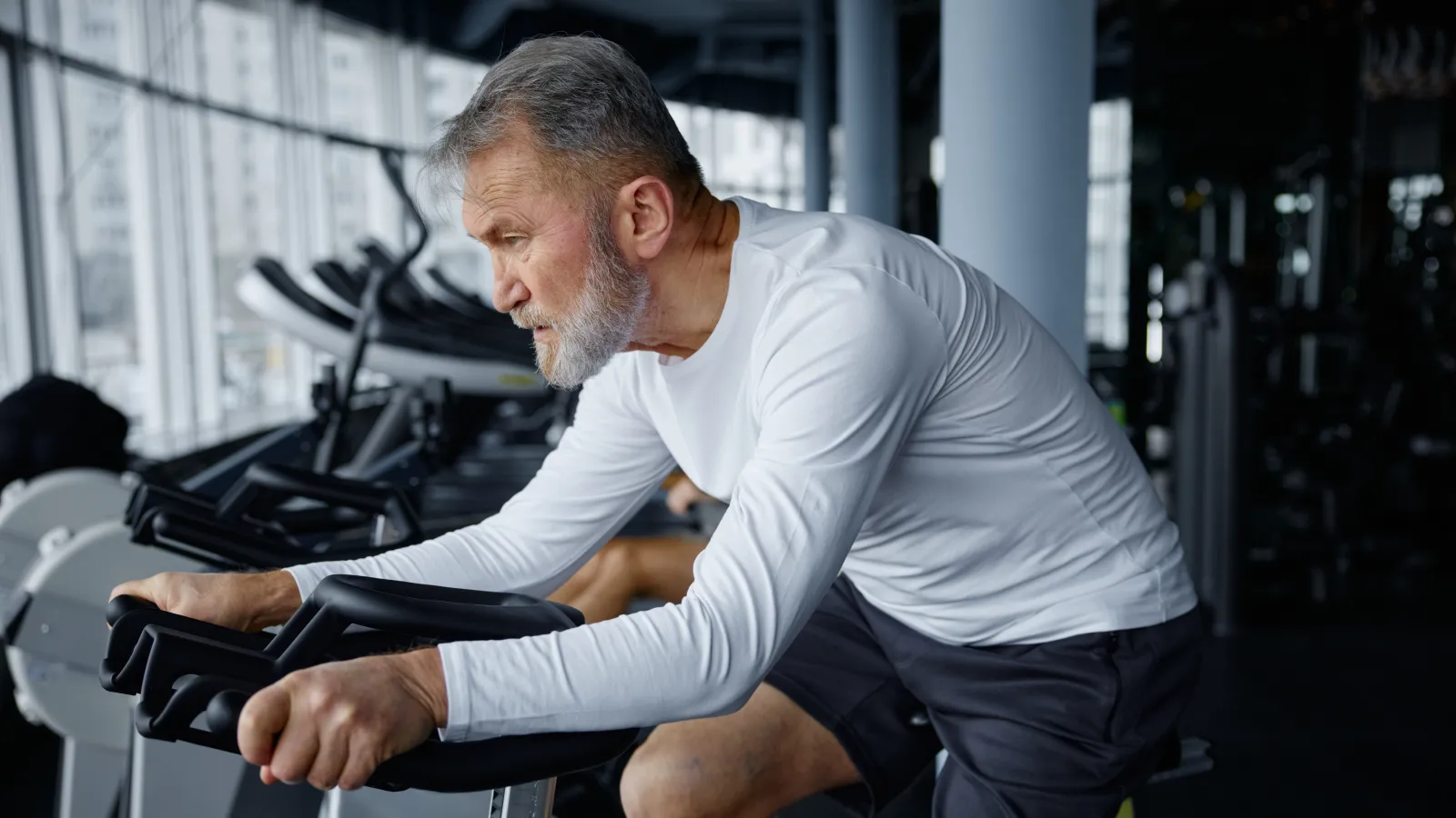 Mature man in white long sleeve shirt exercising on stationary bike in a gym with focused expression.