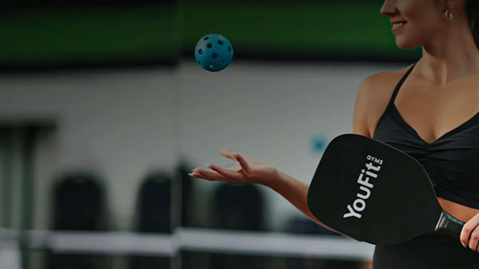 Woman in black workout attire playing pickleball with YouFit paddle and blue ball in indoor gym