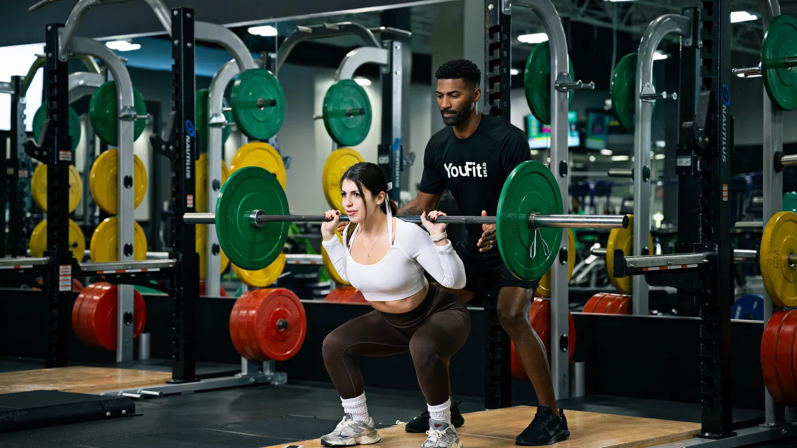 Woman performing barbell squats with trainer assisting in a gym with colorful weight plates.