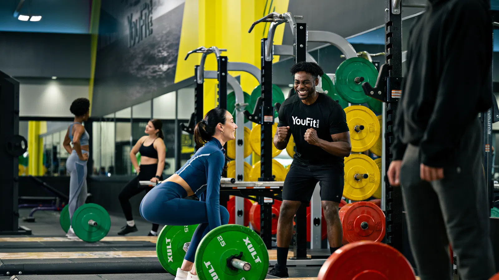 Woman lifting green barbell weights during training session in gym with coach motivating her.