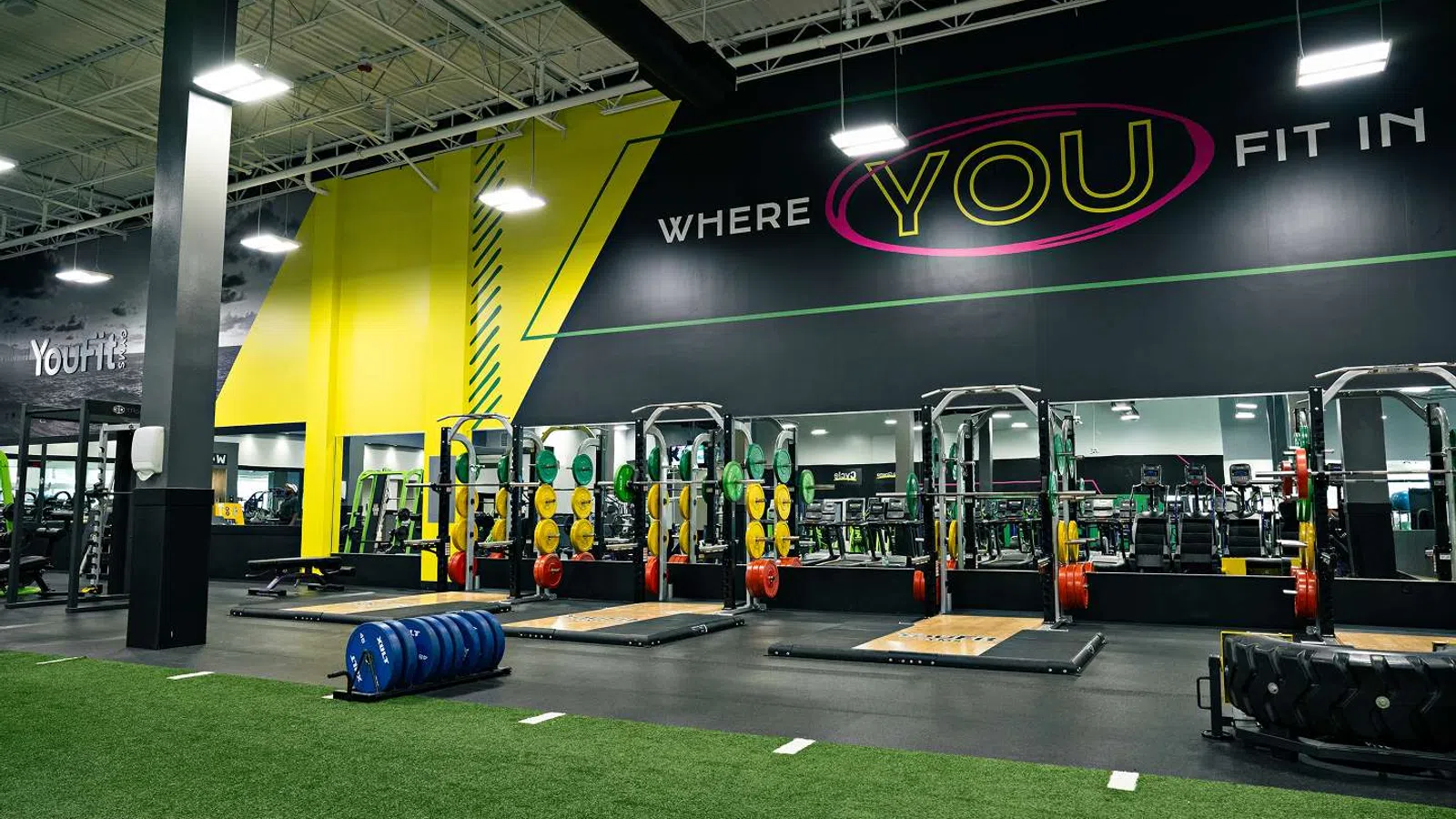 Modern gym interior with weightlifting platforms, colorful plates, turf area, and motivational wall text