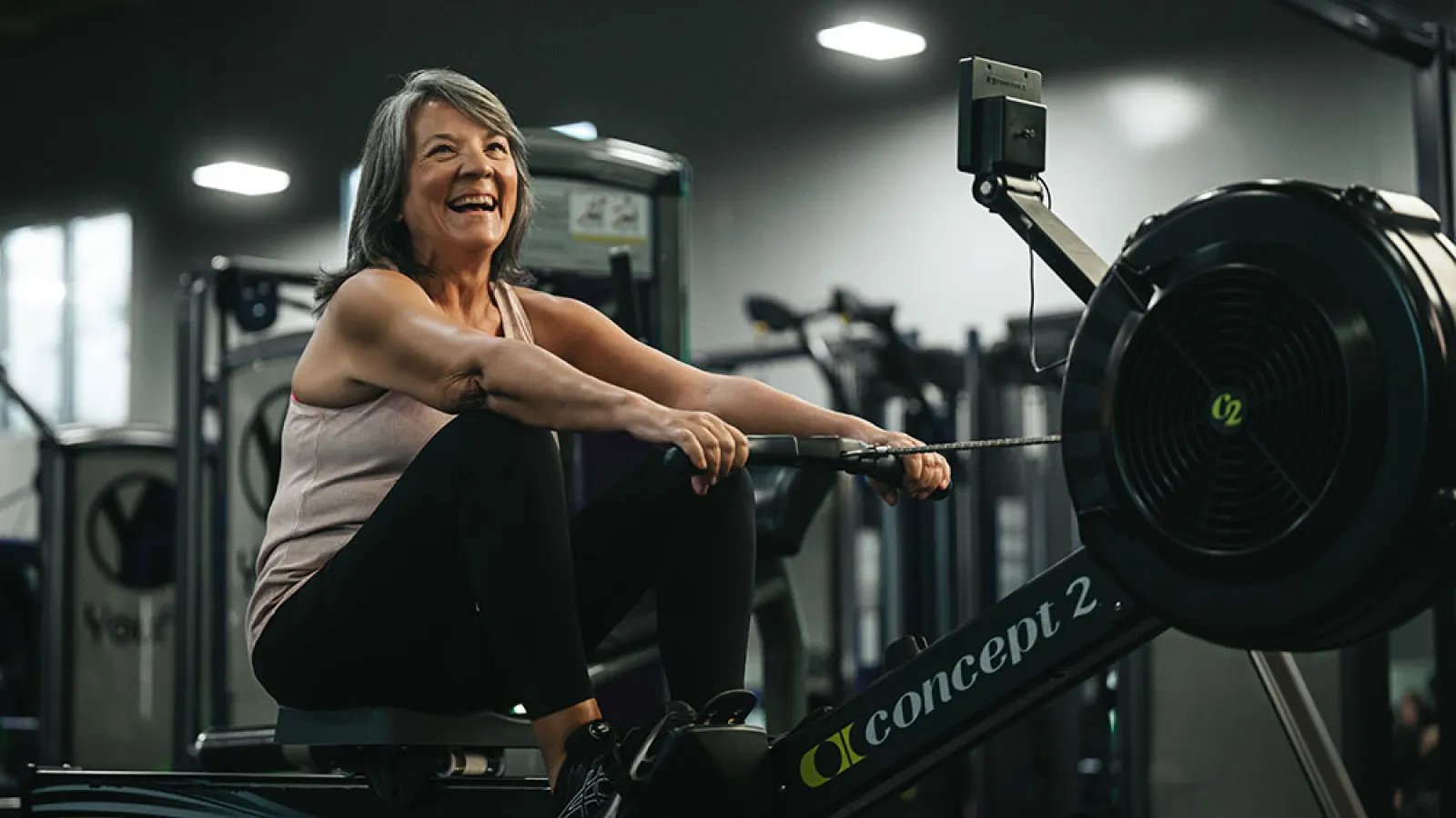 Smiling middle-aged woman using a Concept2 rowing machine in a gym setting, engaging in cardio exercise.
