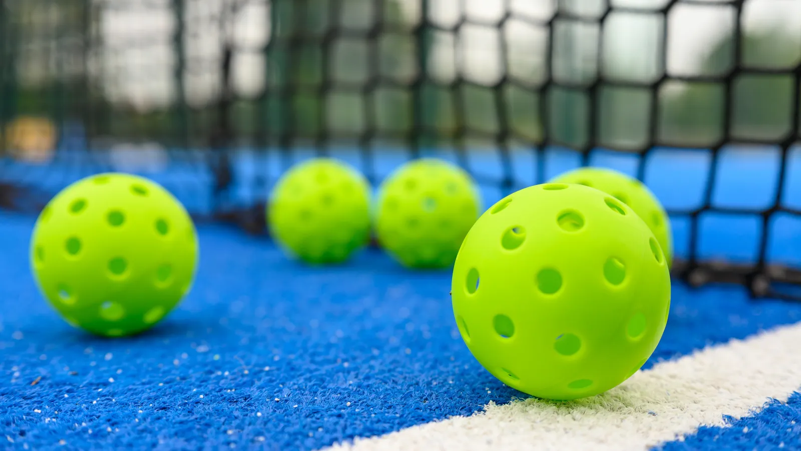 Bright green pickleball balls on a blue court near a black net with white boundary line in focus.