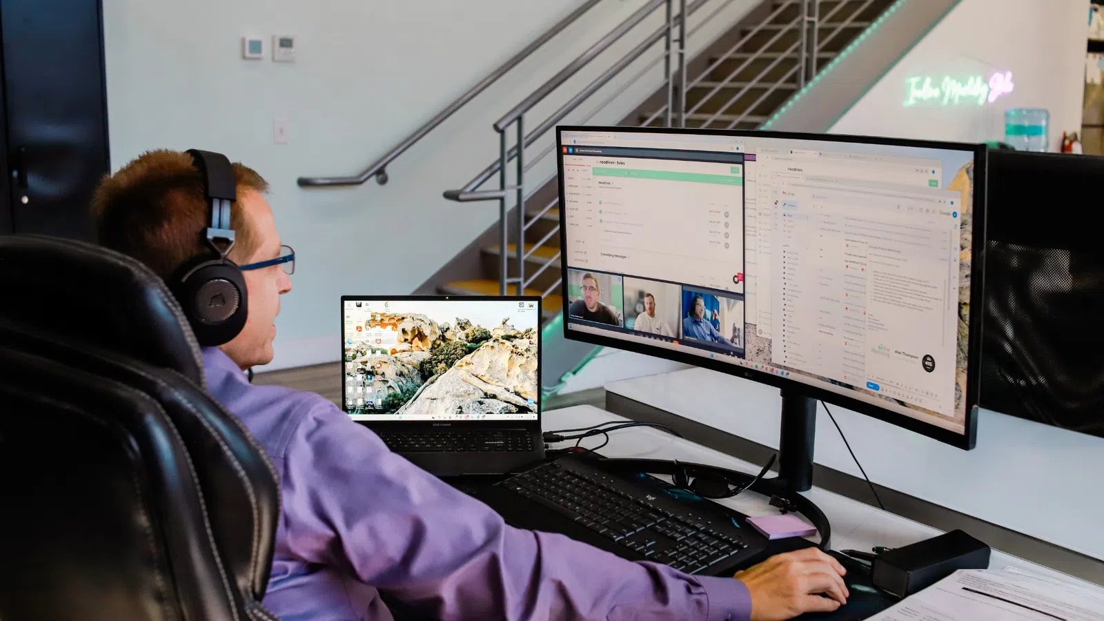 Man wearing headphones working on laptop and dual monitor setup in modern office with staircase in background